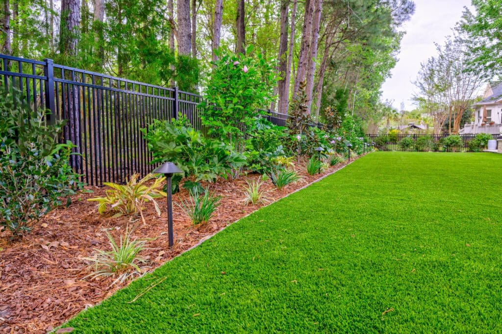 Naturalistic Lowcountry planting border with ferns, ornamental grasses, and shrubs edging a real-grass lawn against a backdrop of native pines